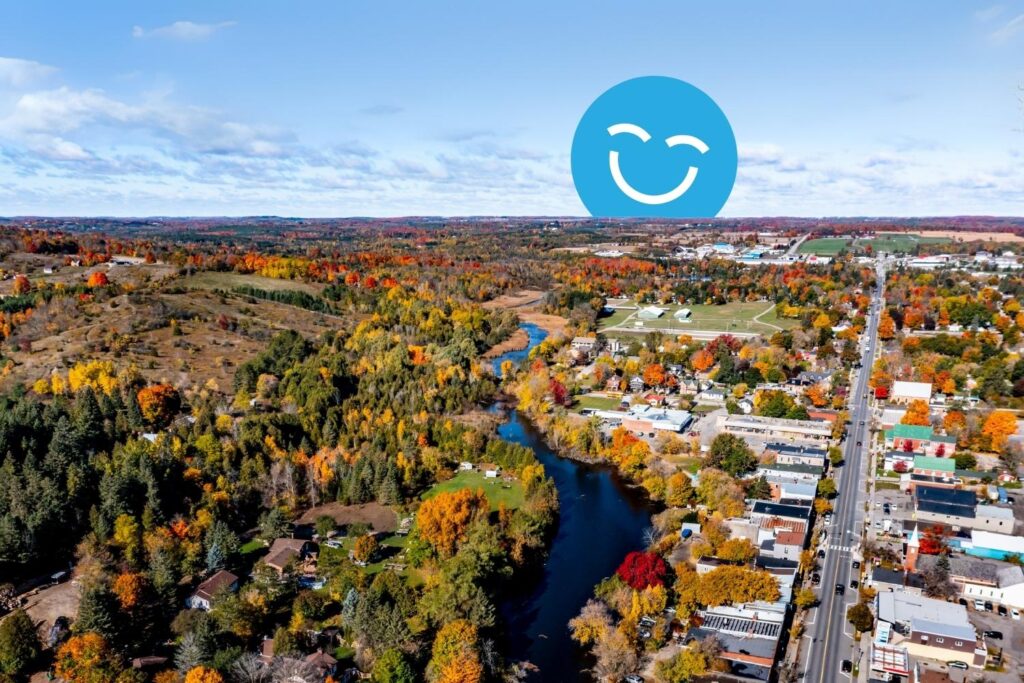 Aerial view of a colorful autumn landscape featuring a winding river through vibrant foliage, with a small town visible along the riverbank. A cheerful blue smiley face icon is superimposed in the sky above.