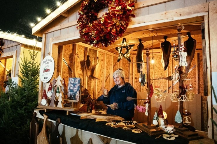 A cheerful vendor stands behind a wooden stall adorned with handmade crafts and decorations. A festive wreath hangs above, and various ornaments are displayed on the table, capturing the spirit of a holiday market. Warm lights illuminate the cozy atmosphere.