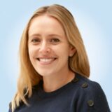 A smiling woman with long blonde hair wears a dark sweater. She has a friendly expression against a soft blue background.