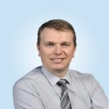 A professional man smiles confidently while wearing a light blue checkered shirt and a grey tie. The background is a soft blue, creating a welcoming atmosphere.