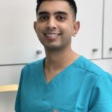 A smiling male healthcare professional wearing a teal scrubs top, standing against a light-colored background.