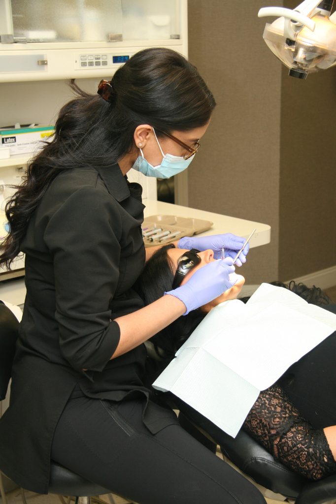 A dental professional wearing a mask and gloves attentively treats a patient reclining in a dental chair. The patient is covered with a dental bib, and dental instruments are visible in the background, highlighting a care-focused environment.