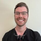 A smiling young adult male with glasses and short hair, dressed in a black shirt. He appears friendly and approachable, standing against a plain light background.