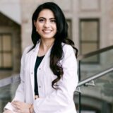 A smiling woman wearing a white lab coat stands confidently near a staircase in a modern building, showcasing a professional and welcoming demeanor.