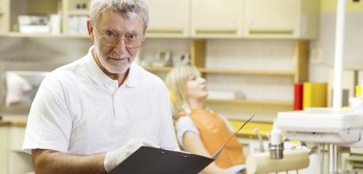 A dentist stands in an office holding a clipboard, looking directly at the camera. In the background, a patient is seated in a dental chair, wearing a bib and appearing relaxed. The setting suggests a routine dental visit.
