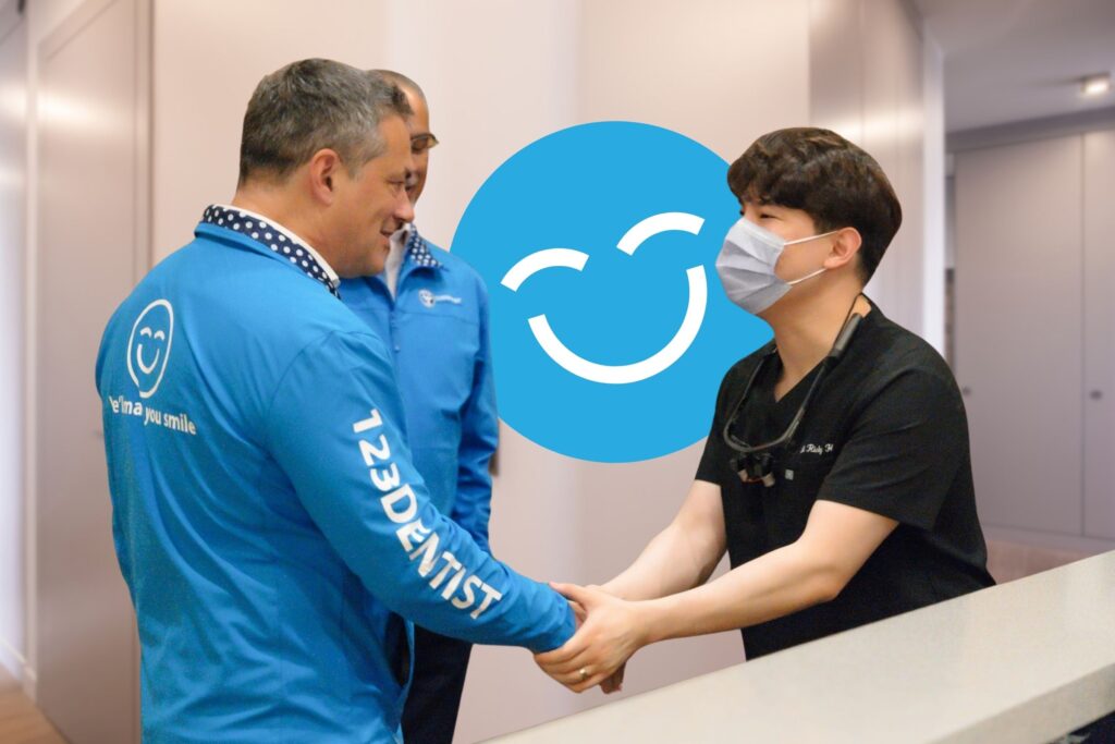 Two people, one in a blue shirt and the other in black, are shaking hands in a dental clinic. The friendly 123Dentist Smiley is prominently featured in the background, adding a cheerful atmosphere to the scene.