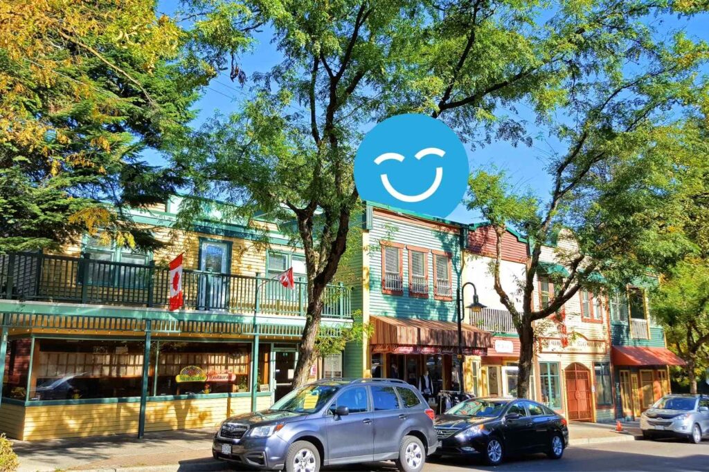 A vibrant street scene featuring colorful buildings with balconies, trees providing shade, and parked cars along the curb, all under a clear blue sky with a smiling face graphic overlay.