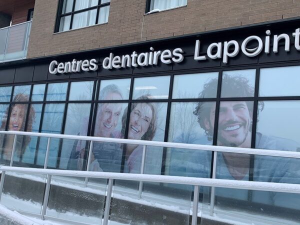A dental clinic storefront featuring large, colorful images of smiling individuals. The sign reads "Centres dentaires Lapointe," and the building is surrounded by snow, indicating a winter setting.