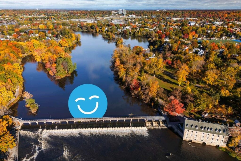 Aerial view of a river with colorful autumn trees lining the shore. A dam is visible on the left, and a blue smiley face graphic is superimposed in the center. The background features a town and a clear sky.