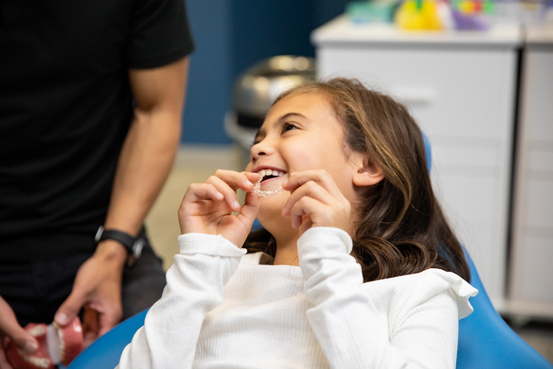 A young girl smiles joyfully while sitting in a dental chair, showcasing her excitement. A dental professional is nearby, attending to her. The setting is bright and friendly, emphasizing a positive dental experience. The 123Dentist Smiley can be seen, adding to the cheerful atmosphere.
