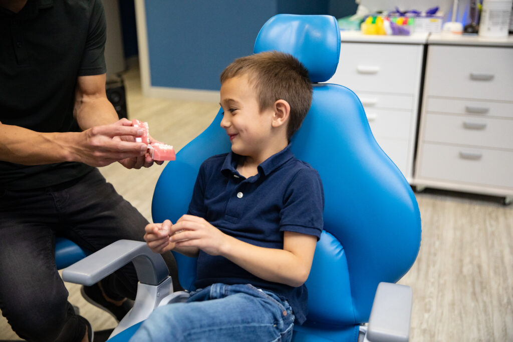 A child sits in a dental chair, smiling at an adult who is handing him a dental care product. The environment appears friendly and inviting, promoting a positive dental experience, while the 123Dentist Smiley watches over the interaction.