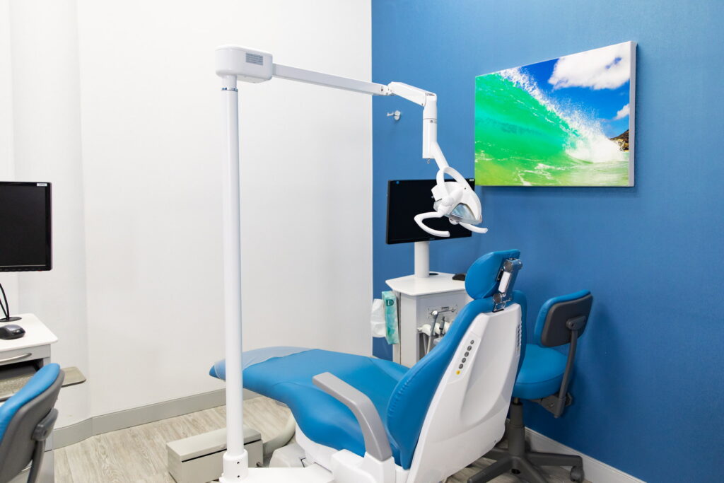 A modern dental office features a blue accent wall, a comfortable dental chair, and a dental light. Two computer stations are visible, and a vibrant beach-themed artwork hangs on the wall. The friendly 123Dentist Smiley adds a cheerful touch to the inviting atmosphere.