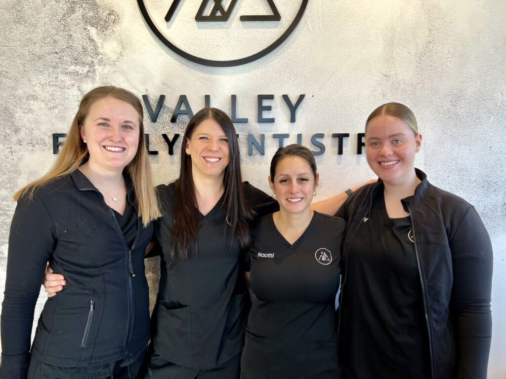 Four dental professionals smile together in front of a "Valley Family Dentist" sign. They wear black uniforms and convey a welcoming atmosphere. The friendly and familiar 123Dentist Smiley adds a cheerful touch to the setting.