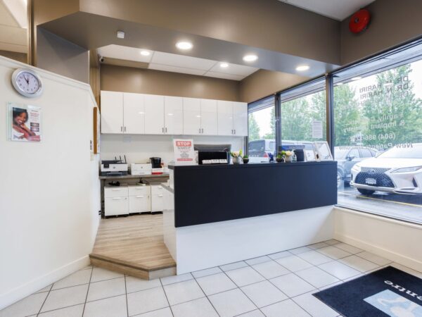 A modern reception area featuring a black front desk, white cabinets, and large windows allowing natural light. A clock and a poster are visible on the walls, with a tiled floor and a welcome mat at the entrance.