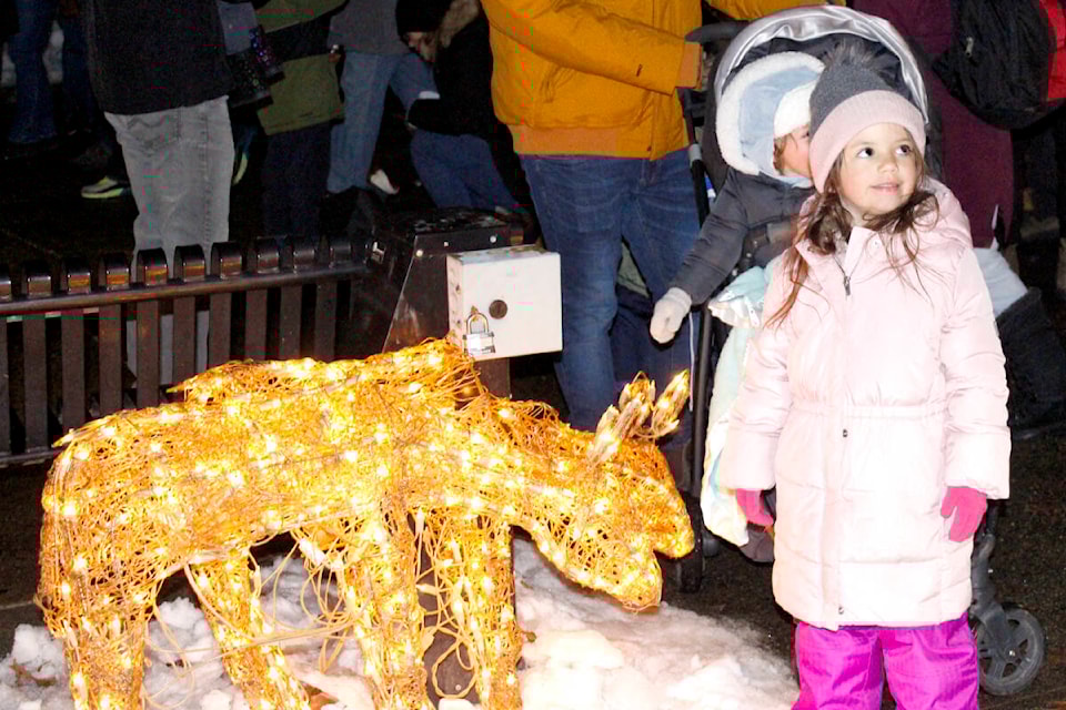 A young girl in a pink coat stands next to a glowing, decorated deer sculpture in a snowy setting, surrounded by people celebrating the festive season.