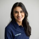 A smiling woman with long dark hair is wearing a navy blue shirt with "Dr. Maria Syed" embroidered on it. She has a friendly expression and is posed against a light background.