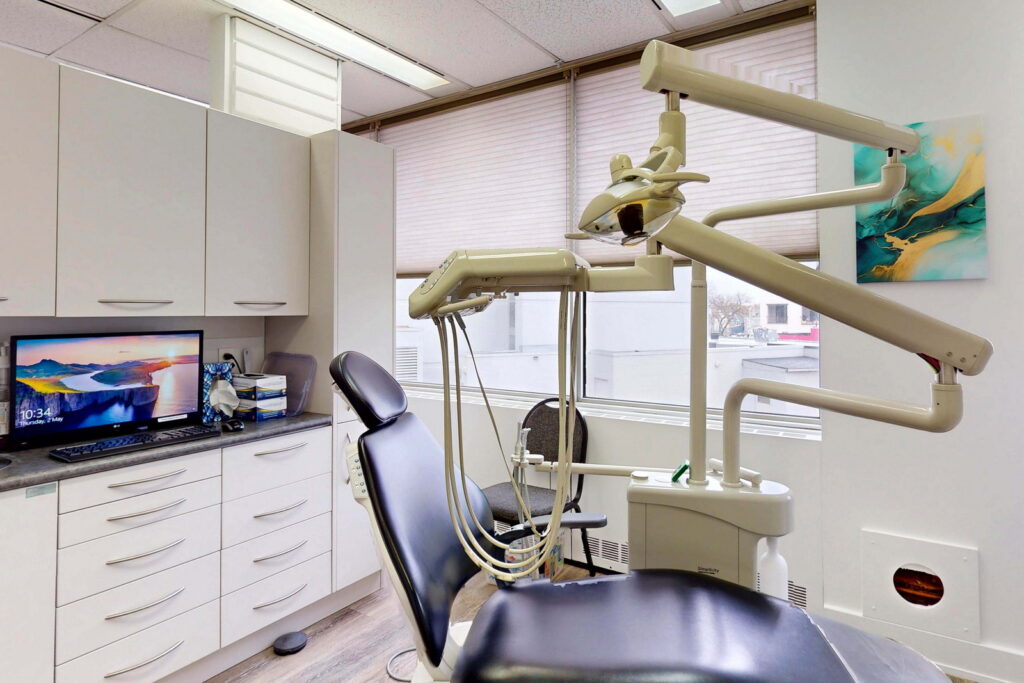 A modern dental office featuring a dental chair, overhead light, and a computer monitor displaying artwork. The space is well-lit with windows and has cabinets for storage, creating a clean and professional atmosphere.