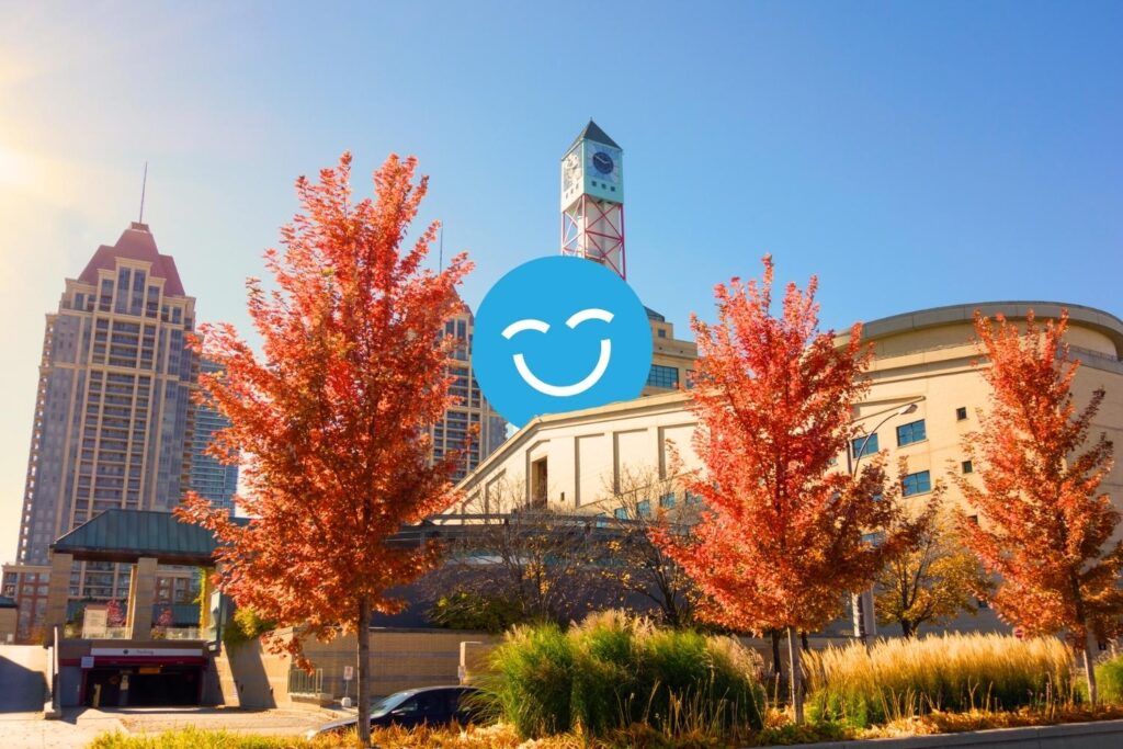 A sunny urban scene featuring modern buildings and vibrant red trees in autumn. A cheerful blue smiley face is superimposed over the landscape, adding a whimsical touch to the cityscape.