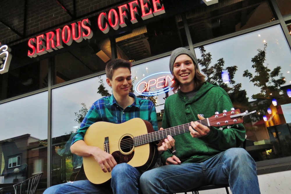 Two young men sit outside a coffee shop, one playing an acoustic guitar while both smile and enjoy the moment. The shop's sign reads "SERIOUS COFFEE," and an "OPEN" sign is visible in the background.