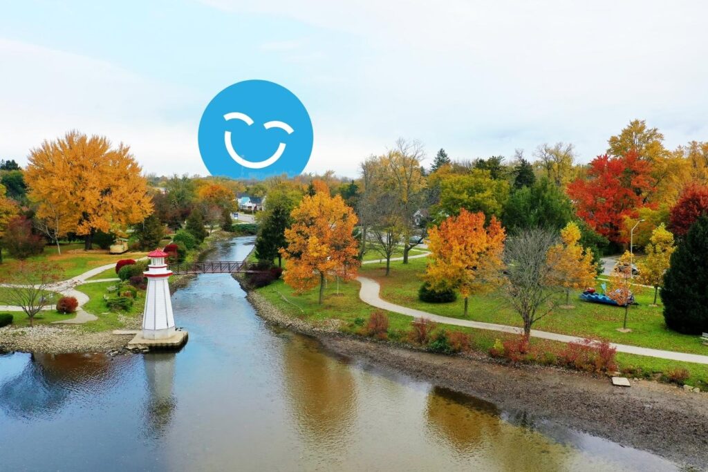 A serene park scene featuring vibrant autumn foliage, a calm river, and walking paths. A white lighthouse stands near the water, complemented by a playful blue smiley face in the background. The landscape captures the beauty of fall in a tranquil setting.