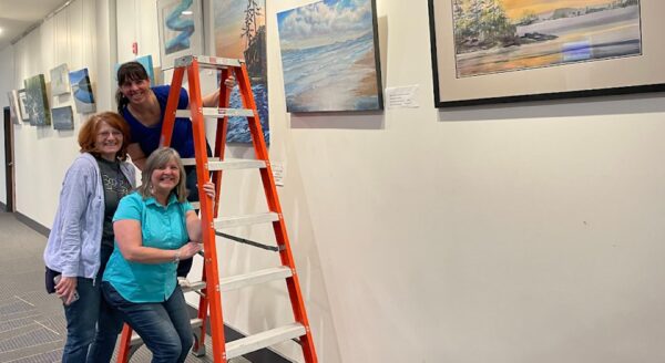 Three women smile in an art gallery as they arrange paintings on the wall. One woman stands on a ladder, while the others pose beside it. The walls are decorated with various landscapes.