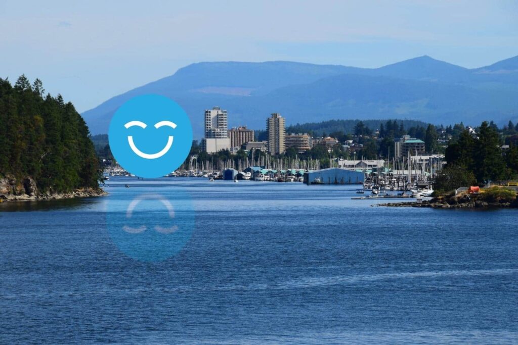 A scenic view of a calm river with lush trees on either side, leading to a city skyline in the background, framed by mountains. A large blue smiley face overlays the image, adding a cheerful element to the landscape.