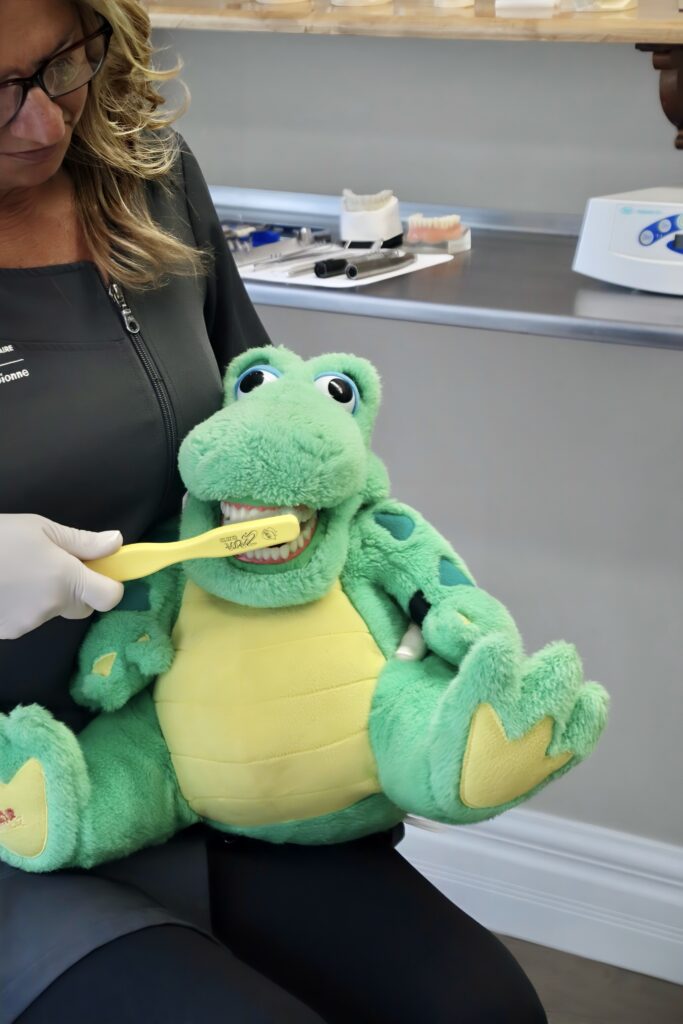 A woman demonstrates proper tooth brushing techniques on a large green stuffed animal shaped like a frog, sitting in a dental office. The frog has its mouth open, and a toothbrush is in use, promoting dental hygiene in a playful manner.