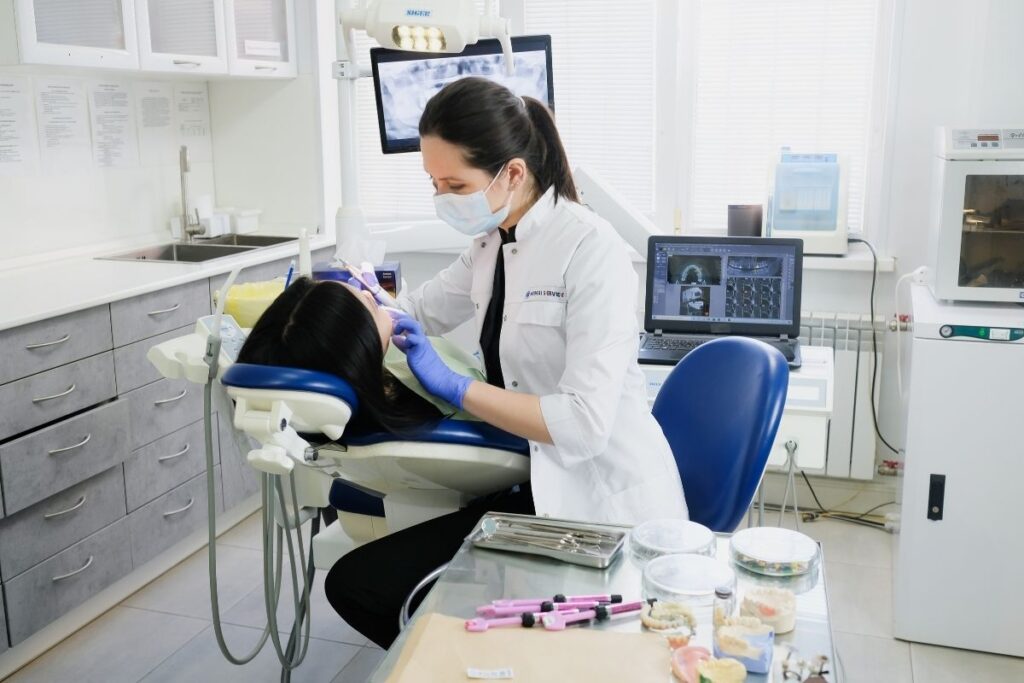 A dentist in a white coat and protective mask examines a patient in a dental chair, using dental tools and technology. The bright, modern clinic is equipped with screens and various instruments, creating an atmosphere of professionalism and care.