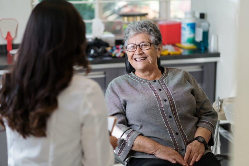 A smiling older woman sits in a dental office, engaging with a dental professional. The setting is bright and welcoming, reflecting a friendly atmosphere. The 123Dentist Smiley adds a cheerful touch to the scene, promoting a positive dental experience.