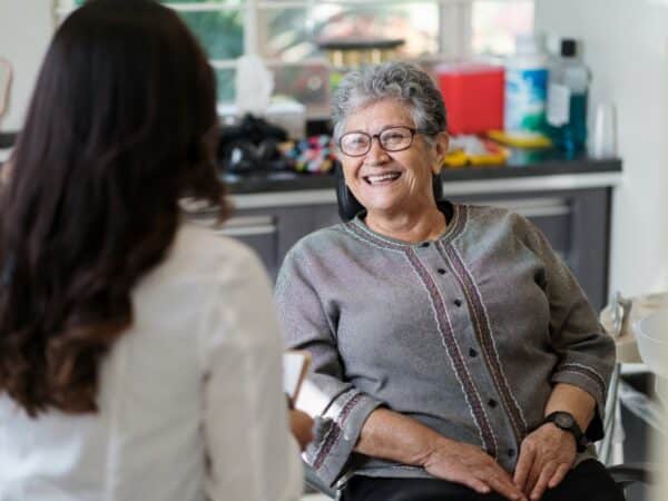 A smiling older woman sits in a dental office, engaging with a dental professional. The setting is bright and welcoming, reflecting a friendly atmosphere. The 123Dentist Smiley adds a cheerful touch to the scene, promoting a positive dental experience.