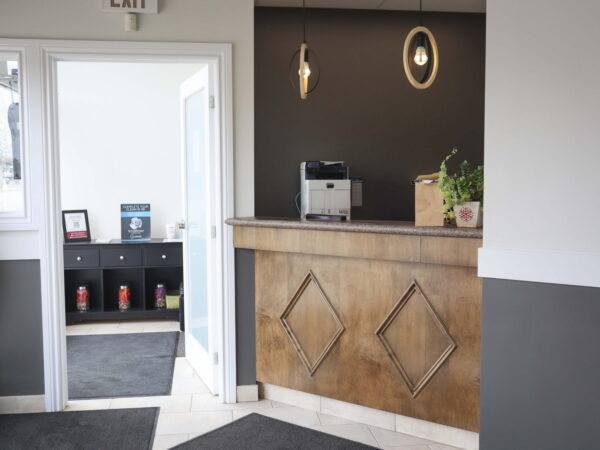 A modern reception area featuring a wooden counter with a diamond design, a coffee maker, and potted plants. The space is well-lit with a door leading to another room and a black wall providing contrast. Stylish lighting fixtures add an inviting atmosphere.