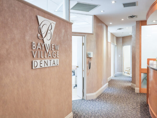 The image shows the interior of a dental office, featuring a sign that reads "Bayview Village Dental." The welcoming reception area has a warm color scheme and leads to a corridor, creating a professional yet friendly atmosphere.