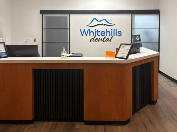A dental office reception area featuring a modern wooden front desk, a logo for "Whitehills Dental" on the wall, and informational displays. The space is well-lit with a clean and inviting aesthetic.