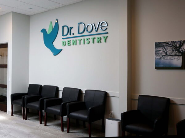 A dental office interior featuring a sign that reads "Dr. Dove Dentistry" alongside a blue dove graphic. There are several black chairs arranged against a light-colored wall, with a framed image of a tree on display.