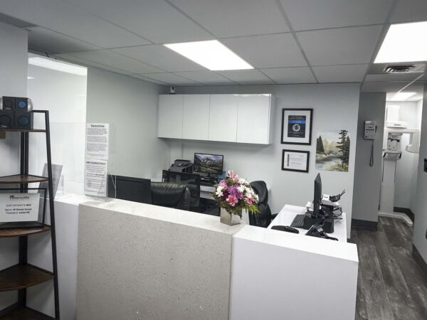 A modern office reception area featuring a low countertop with a flower arrangement, black office chairs, and a wall with framed artworks. Bright overhead lighting illuminates the gray walls and wood-finished flooring, creating a welcoming environment.
