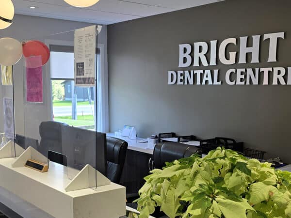 The image shows the interior of the Bright Dental Centre, featuring a reception area with a modern design, plants, and a welcoming atmosphere. 123Dentist Smiley adds a friendly touch to the environment, inviting visitors into a caring dental practice.