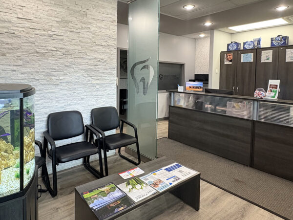 A modern dental office reception area featuring two chairs, a coffee table with magazines, and a fish tank. The front desk is visible, with dental-themed decor. The welcoming atmosphere includes the friendly 123Dentist Smiley, adding a cheerful touch to the space.