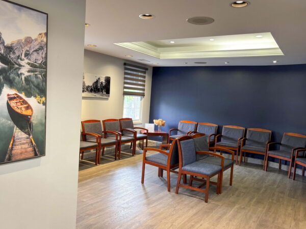 A waiting room featuring wooden chairs arranged in a semi-circle, with a large painting of a boat on water on one wall. The space has natural lighting and a minimalist design, creating a calm atmosphere.
