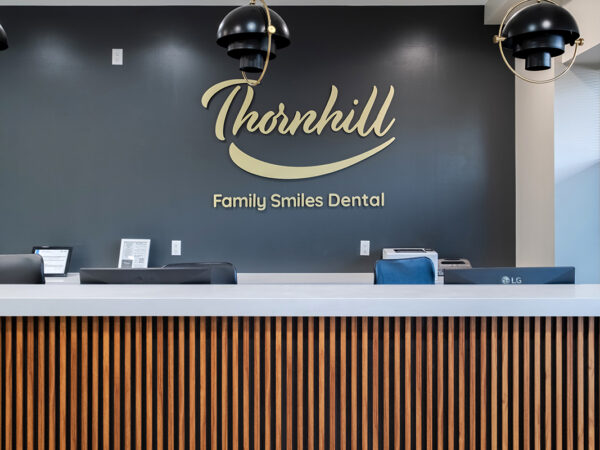 A modern dental reception area features a dark wall with the "Thornhill Family Smiles Dental" sign in stylish lettering. The front desk is sleek, with a light-colored counter and decorative lighting overhead, creating a welcoming atmosphere for patients. The friendly 123Dentist Smiley adds a cheerful touch.