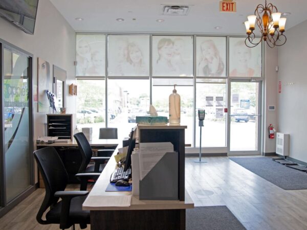 A modern reception area with a wooden desk, black chairs, and large windows showing outdoor light. The space features a neutral color scheme and decorative elements, suggesting a welcoming atmosphere for visitors.