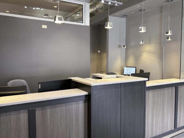 A modern reception area featuring two counter spaces, sleek wooden finishes, and minimalistic design. Soft lighting and technology are present, creating an inviting atmosphere for visitors.