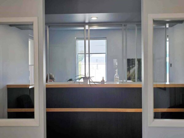 A modern reception area featuring a low wooden counter, framed by glass panels, set against a light gray wall. A window provides natural light, and the space appears organized and welcoming.