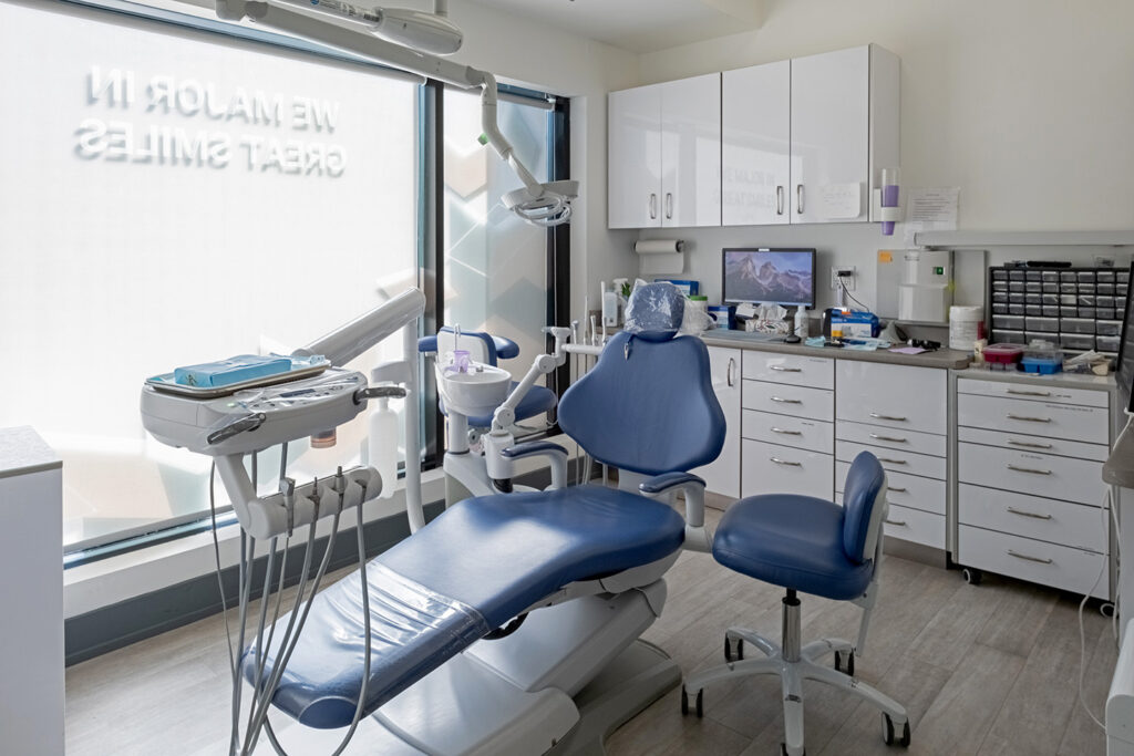 A modern dental clinic featuring a blue dental chair, medical equipment, and a sunny window with the phrase "New Home in Great Smiles" visible. The space is bright, organized, and welcoming, perfect for patient care. The friendly 123Dentist Smiley adds a cheerful touch.