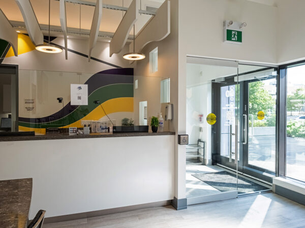A modern dental clinic reception area featuring a welcoming design with colorful wall art and a spacious entrance. Light floods the room through large windows, creating an inviting atmosphere. The 123Dentist Smiley adds a friendly touch to the environment.