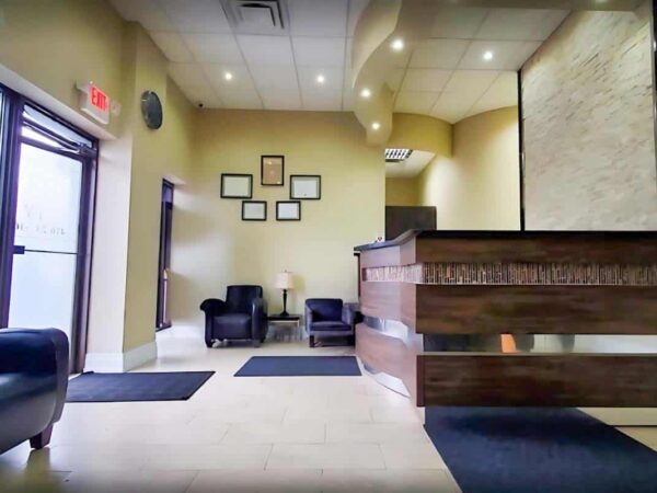 A modern reception area with a wooden front desk, two chairs, and framed artwork on the wall. Natural light enters through a front door, highlighting the clean, welcoming atmosphere.