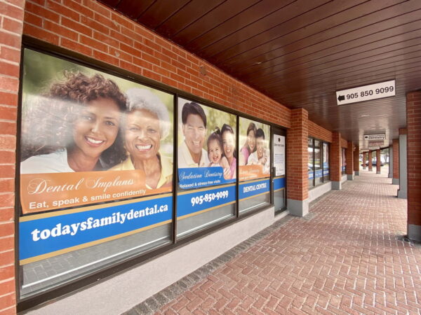 A storefront features promotional windows for a family dental office, showcasing smiling families. The exterior is brick with a covered walkway, and the signage includes contact information and website details, emphasizing dental services for families.