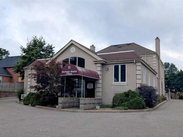 A light-colored building with a sloped roof and an awning, surrounded by trees and bushes, situated on a quiet street. The establishment features large windows and a welcoming entrance, contributing to a friendly atmosphere.