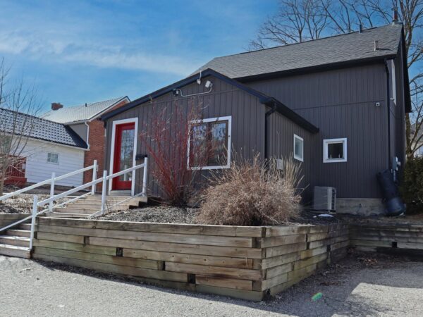 A modern, dark-colored house with a front staircase and wooden landscaping. The house features a red door and large windows, surrounded by minimal vegetation and other residential buildings under a clear blue sky.