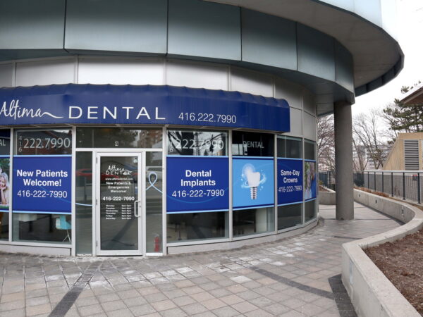 A dental office exterior featuring large windows and signage promoting services like dental implants and welcoming new patients. The building has a modern design with a curved facade, surrounded by paved walkways and landscaping.