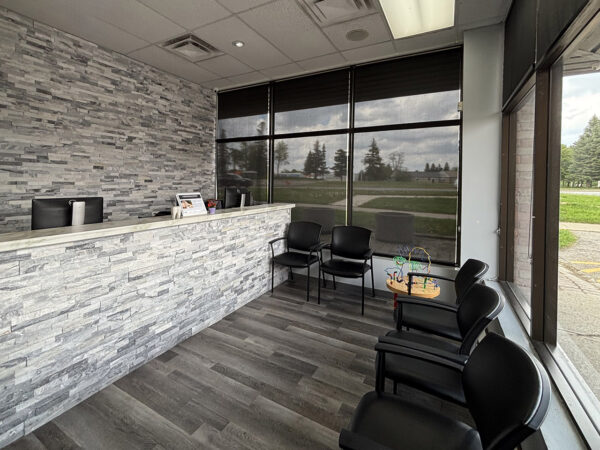 A modern reception area featuring a textured stone front desk, black chairs, and large windows allowing natural light. The flooring is wooden, and there are minimal decorative items on the desk, creating a clean and inviting atmosphere.